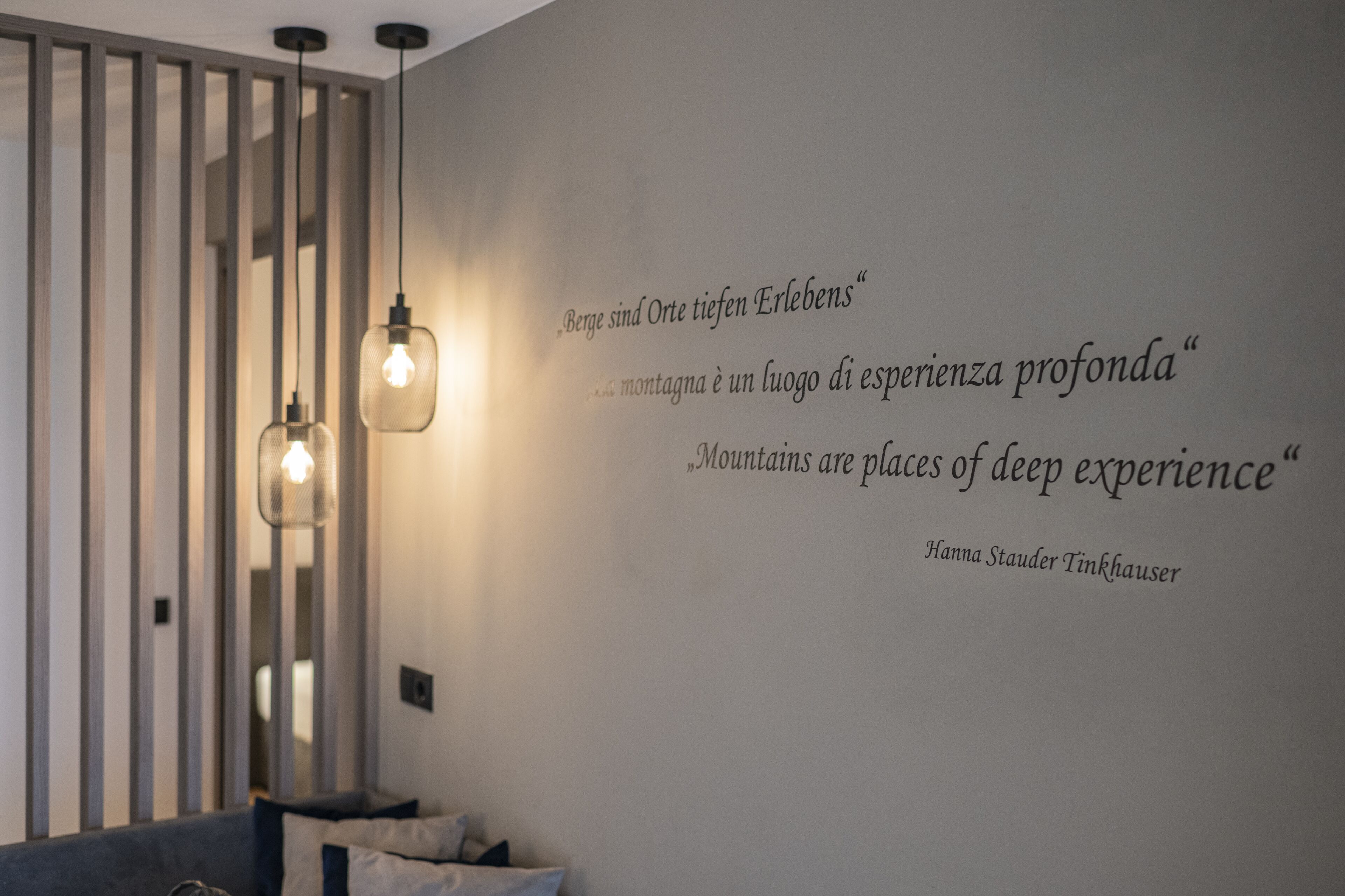 A modern room with a wall-mounted quote by Hanna Staudner Tinkhäuser, a bed, and two hanging pendant lights.