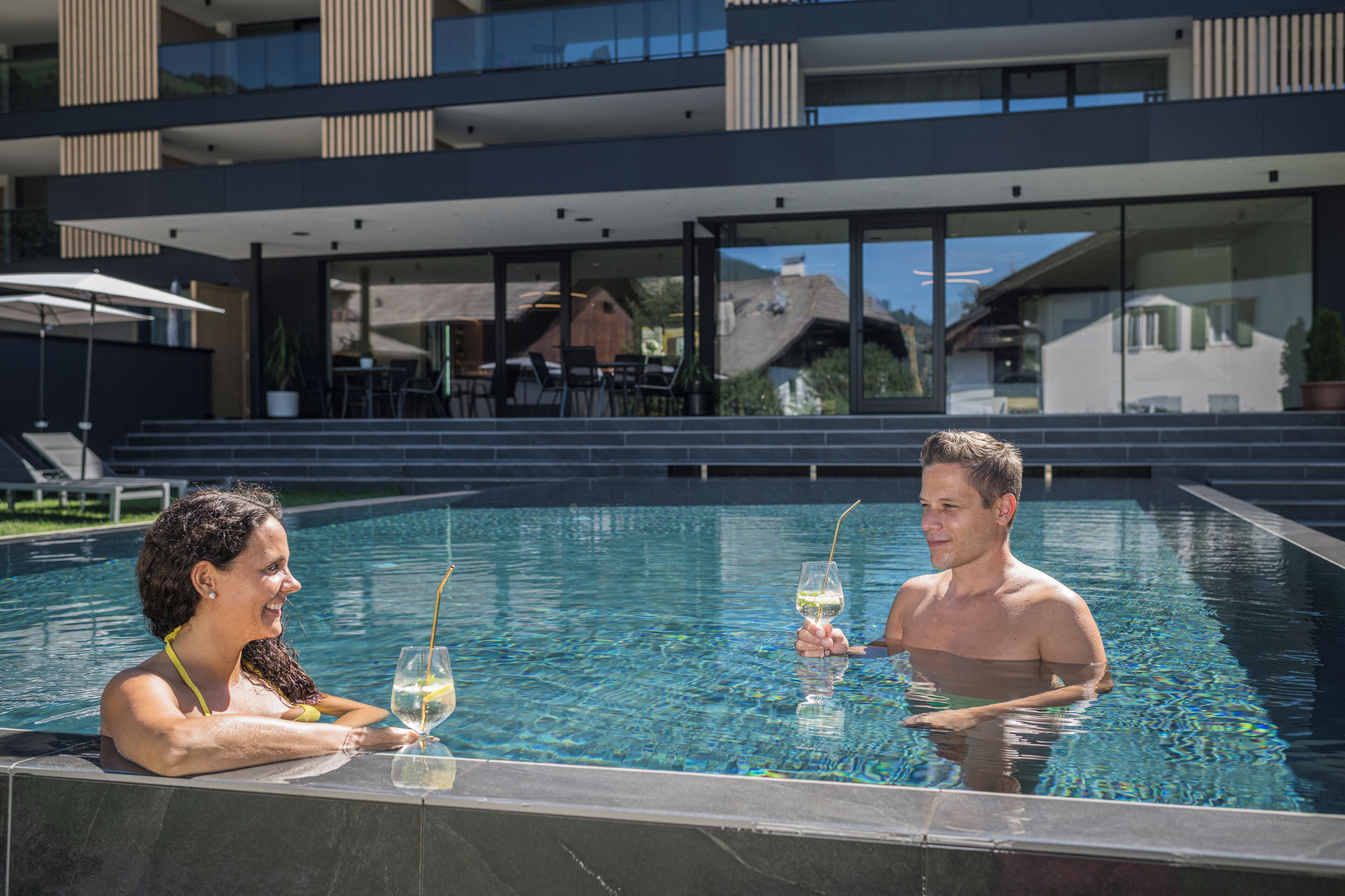 A modern pool area with a glass barrier, a dining area with outdoor seating, and a view of a multi-story building in the background.