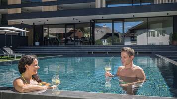 A modern pool area with a glass barrier, a dining area with outdoor seating, and a view of a multi-story building in the background.