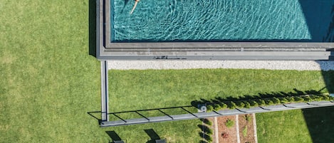 Vue aérienne d’une piscine où une personne flotte, entourée d’une pelouse bien entretenue et de deux tables d’extérieur protégées par des parasols.