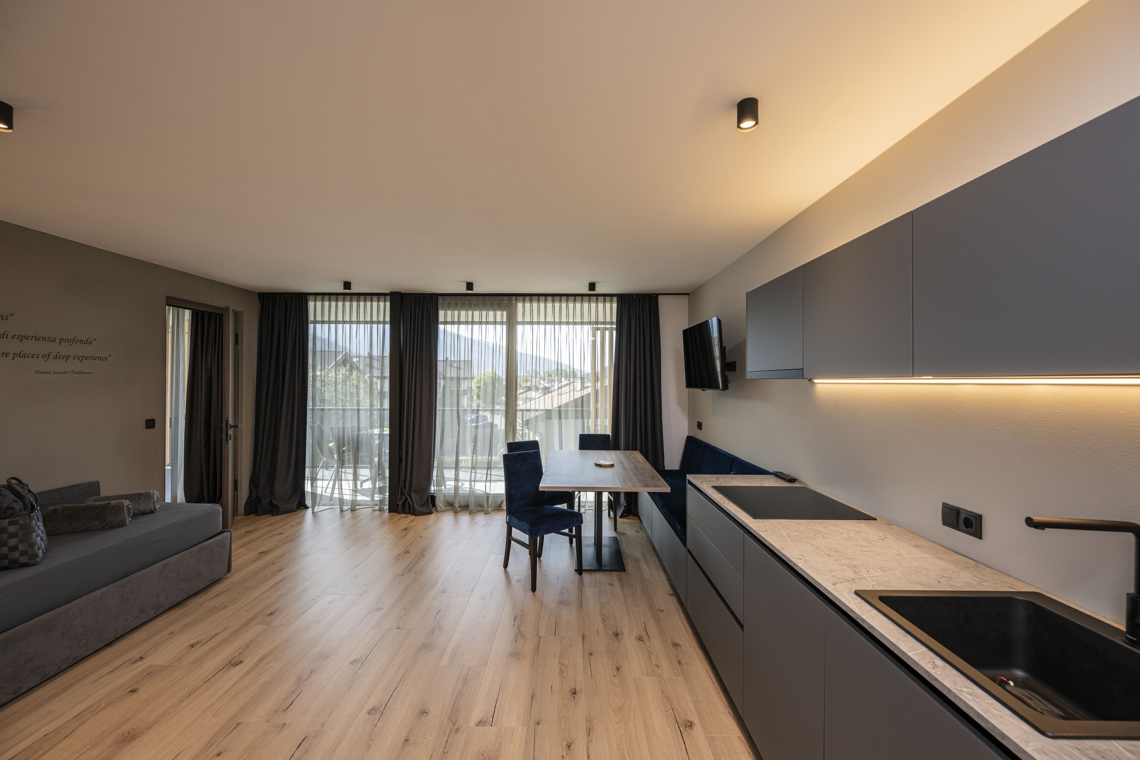 A modern kitchen with a dining area, wooden flooring, and a large window with curtains.