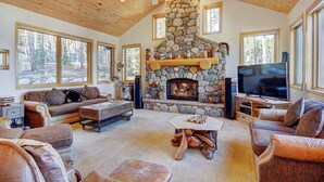 A cozy living room with a stone fireplace, wooden ceiling, and large windows offering a forest view. - Boulder Lodge At Peak 8 4 Bedroom Home by RedAwning (Breckenridge)