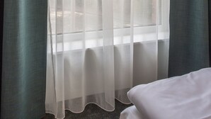 Una habitación con un ventanal grande cubierto por cortinas blancas translúcidas, una cama con ropa blanca y vistas a árboles y un edificio.