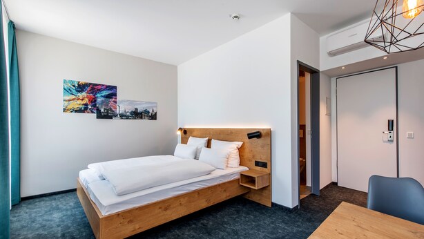 Una habitación de hotel moderna con una cama de madera, un escritorio y una silla. Hay dos cuadros en la pared y una lámpara en el techo.