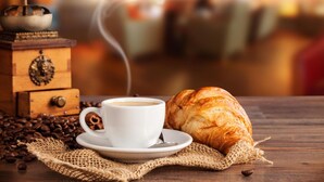 Una taza de café, un croissant y un molinillo de café sobre una mesa de madera con granos de café esparcidos.
