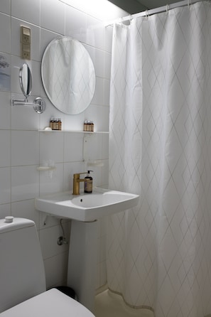 Un baño con lavamanos, espejo e inodoro blancos. Hay repisas con artículos de aseo y una cortina de ducha.