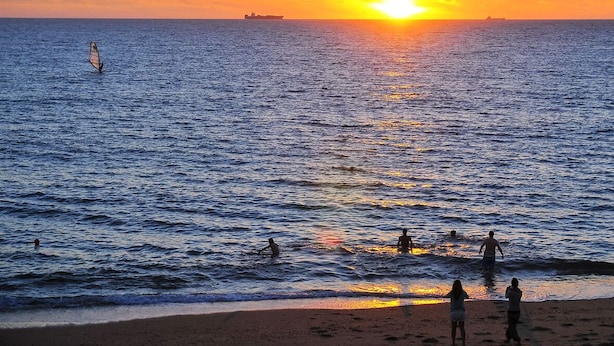 A beach scene with a sunset over the ocean, a sailboat, and people in the water. - National Theater Houses 20 (Porto)