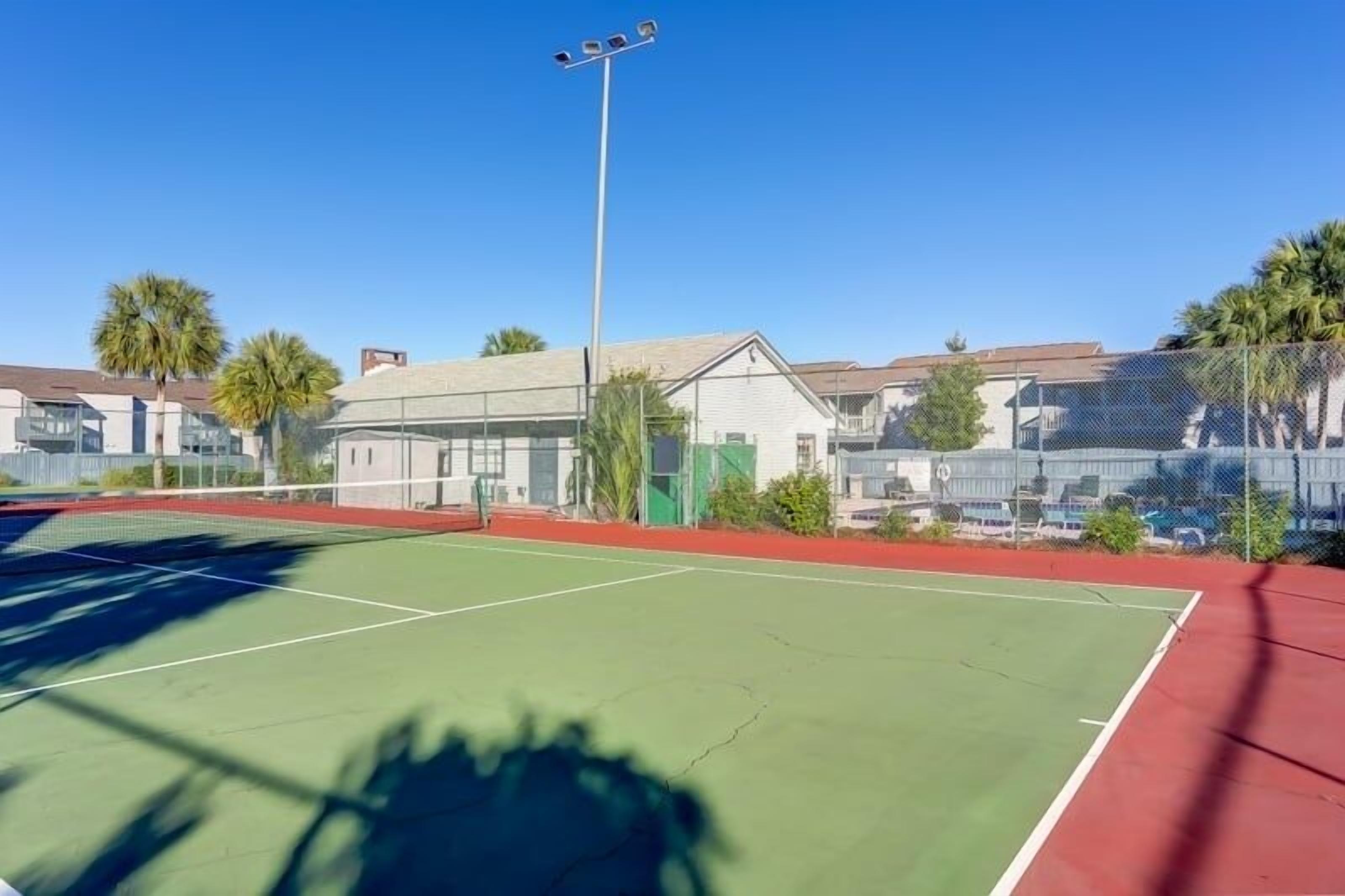 An outdoor tennis court with a net, surrounded by a fence, and adjacent buildings.