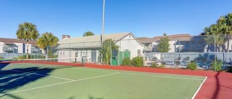 An outdoor tennis court with a net, surrounded by a fence, and adjacent buildings.