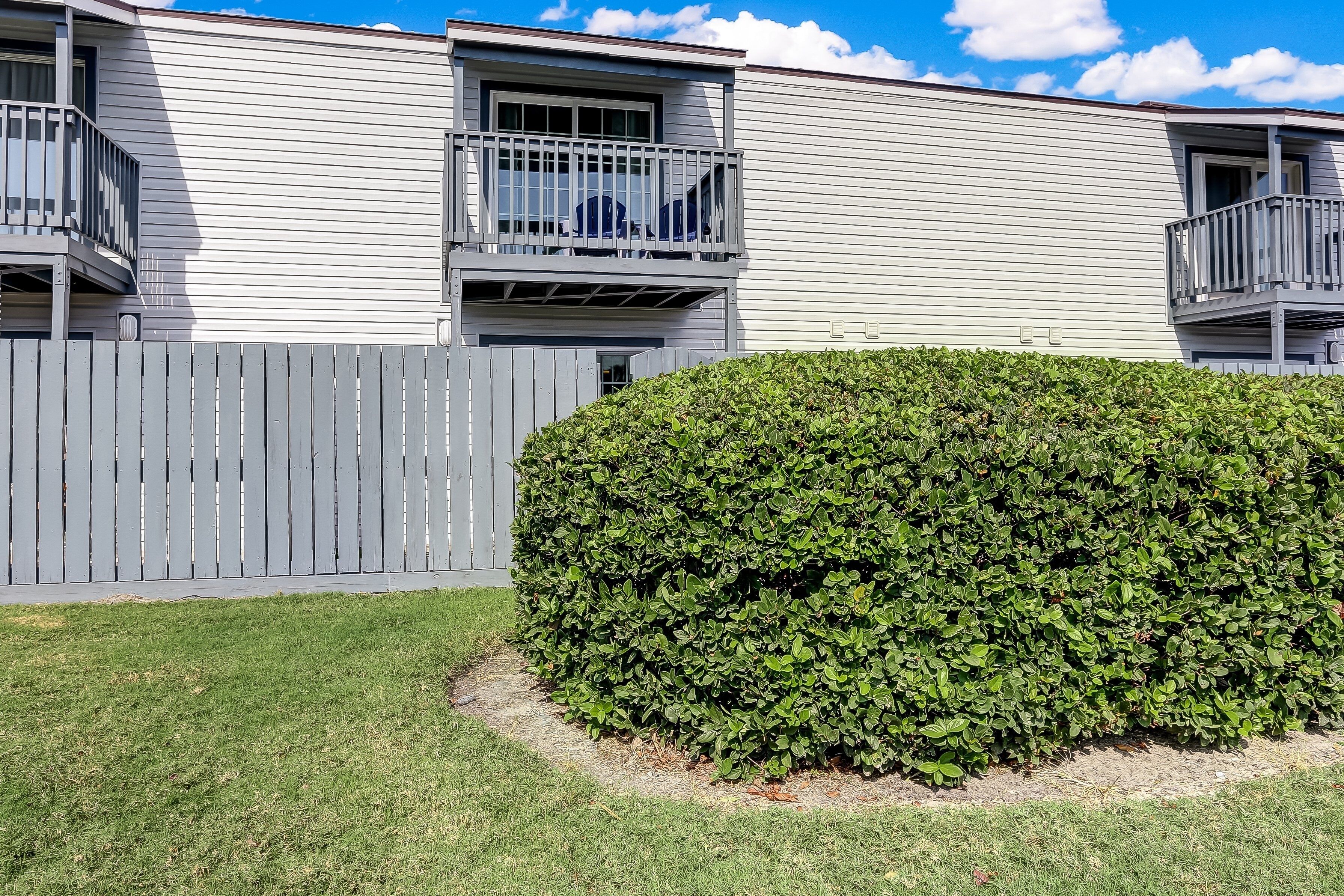 A two-story building with a grey exterior, a wooden fence, and a well-maintained hedge.