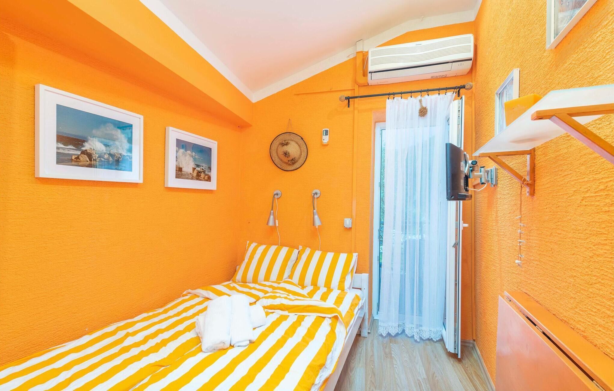 A small, bright room with orange walls, a bed with striped bedding, a ceiling fan, and a window with a curtain.