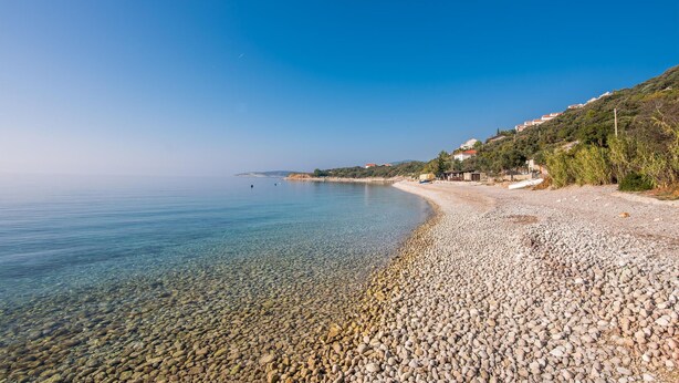 Ein Kiesstrand mit klarem Wasser, einigen Booten und Gebäuden an einem Hügelhang.
