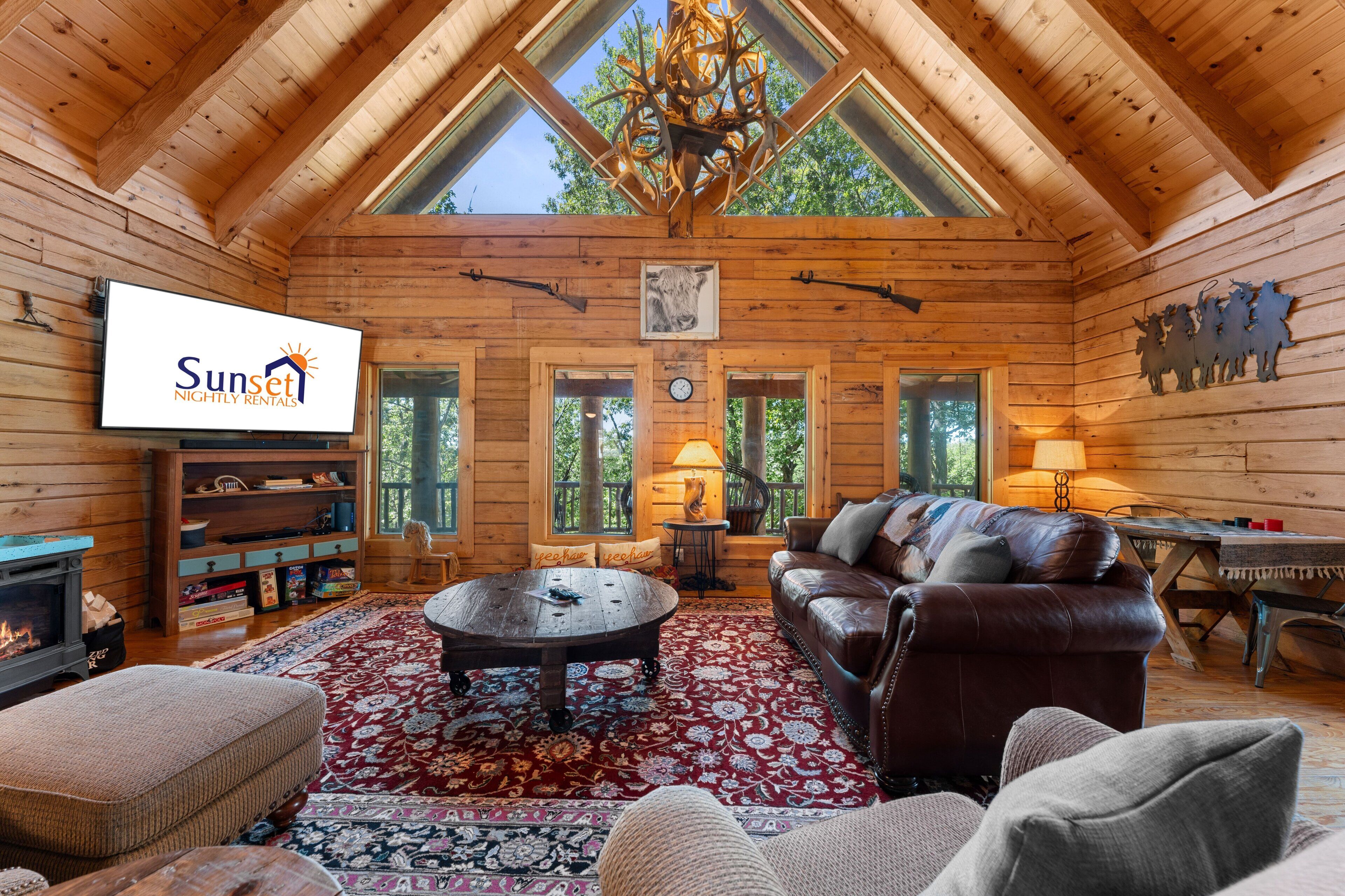 L’intérieur d’une cabane en rondins, avec une cheminée, un canapé en cuir, des murs en bois et un téléviseur diffusant des images de « Sunset Nightly Rentals ».