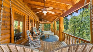 Une cabane en rondins de bois dotée d’un porche comprenant des fauteuils en osier, une table et un ventilateur de plafond.