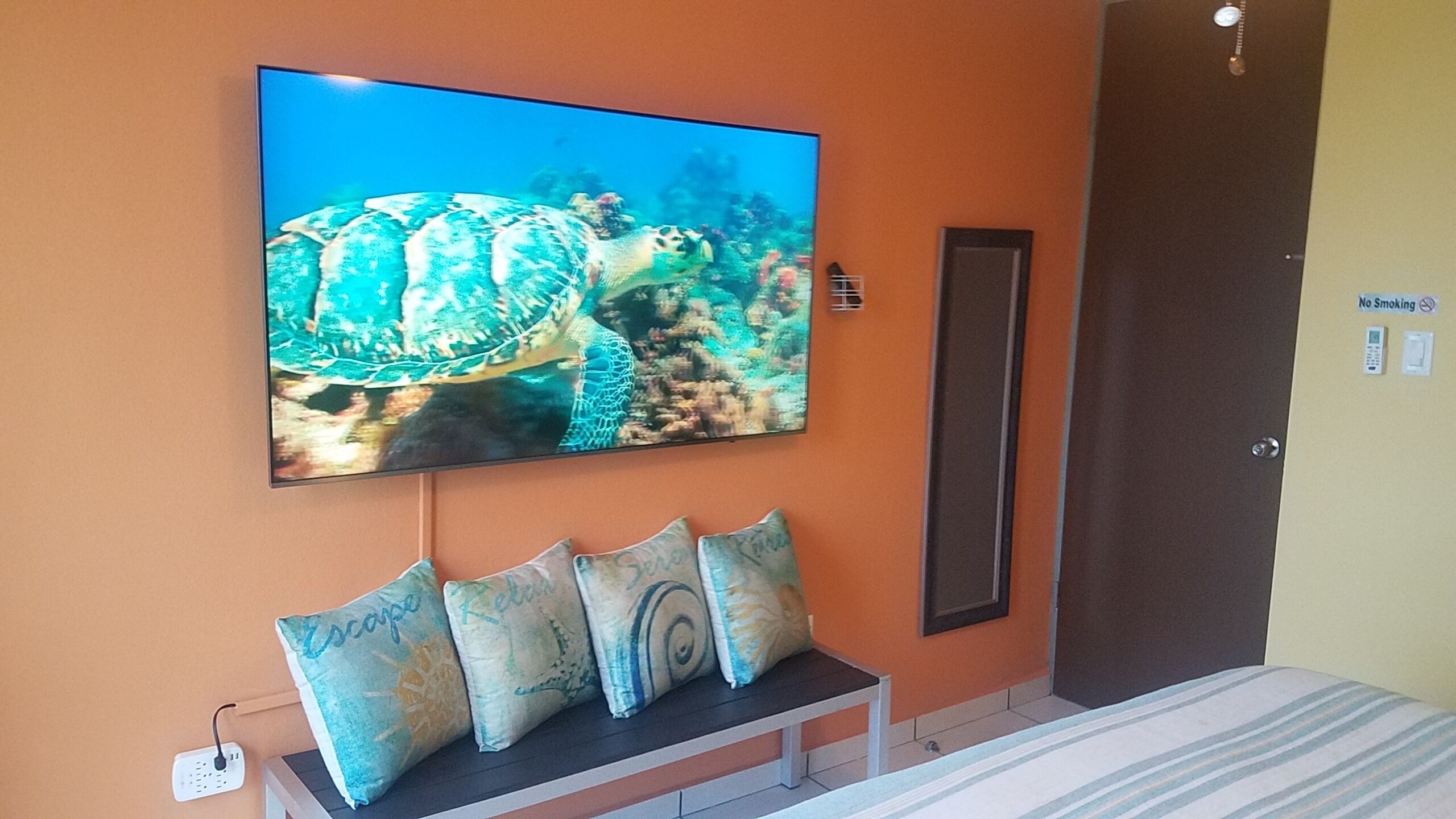 A hotel room with a large TV mounted on the wall displaying an underwater scene, a bench with decorative pillows, and a door with a 'No Smoking' sign.