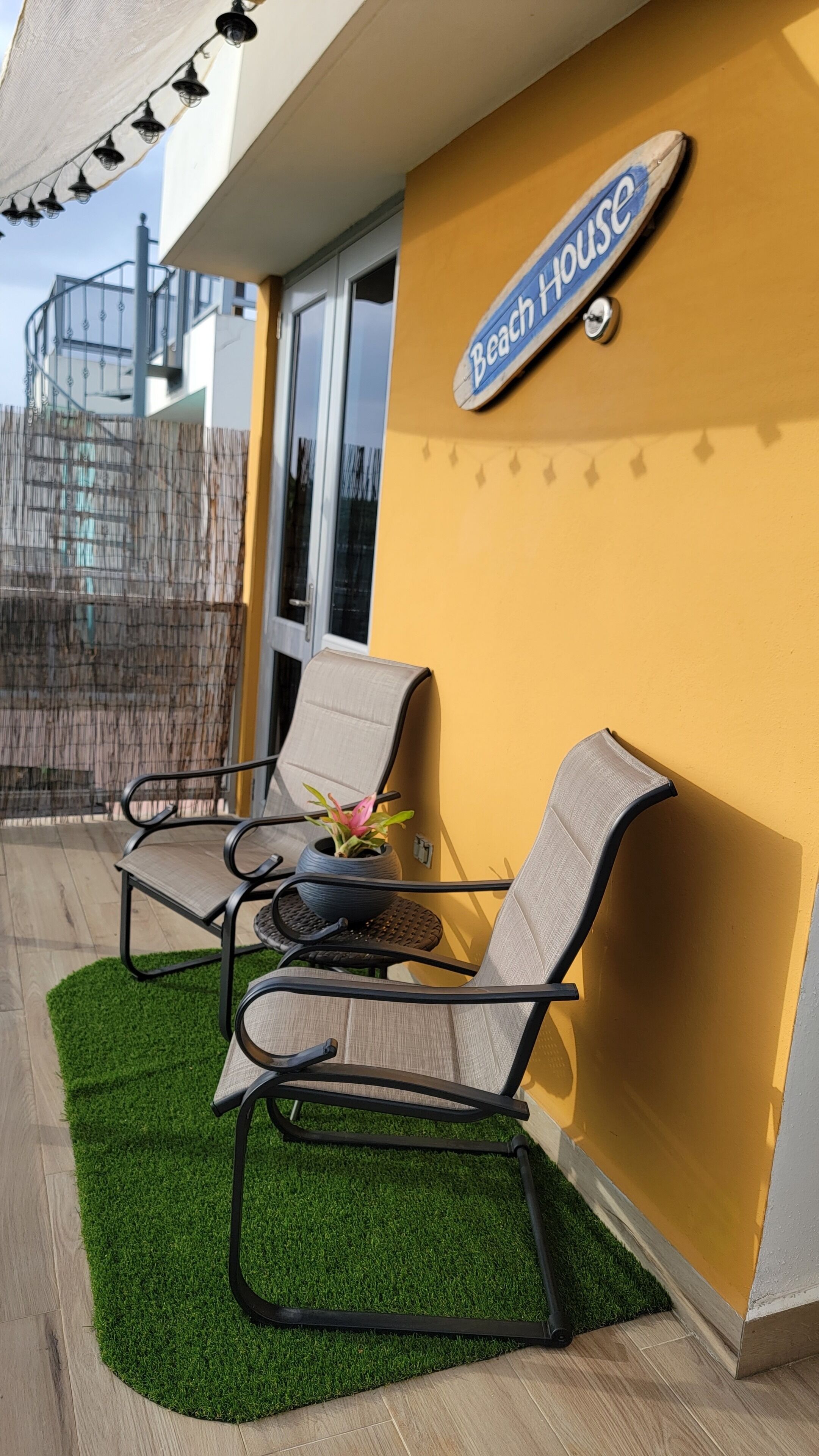 A balcony with two chairs and a small table, a 'Beach House' sign, and a green artificial turf.