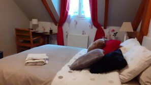 Een slaapkamer met een bed, kussens, een stoel, een tafel en een raam met gordijnen.