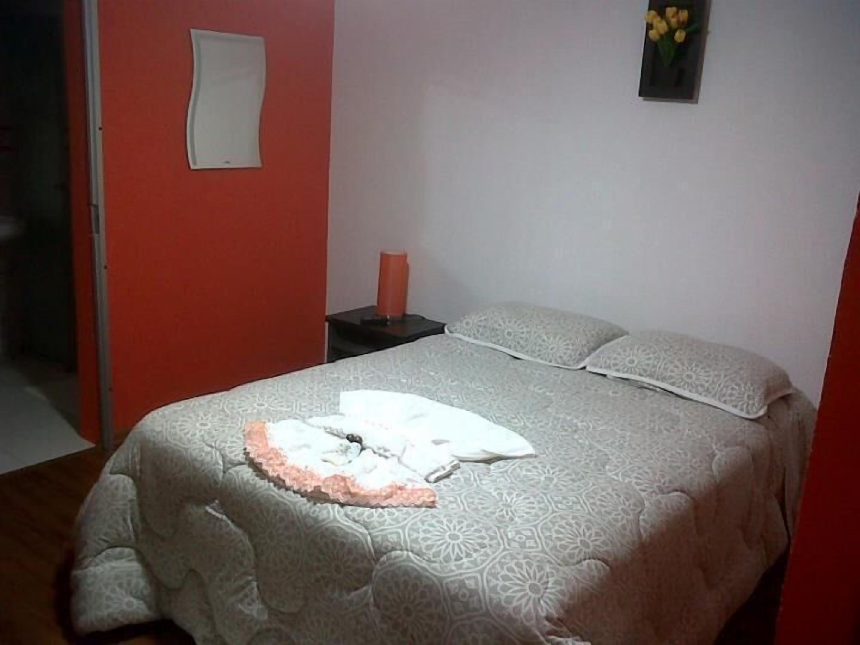 Quarto com cama, mesa de cabeceira, um vaso com flores e parede vermelha.