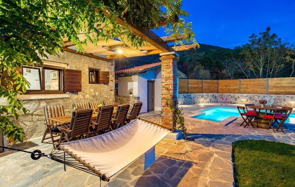 Relax in style on this patio, complete with a dining table, chairs, and hammock, overlooking a swimming pool and lush surroundings.