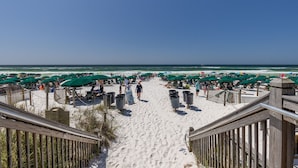 Una spiaggia con una passerella in legno che conduce alla sabbia, fiancheggiata da ombrelloni verdi e sdraie.