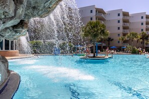 Ein Schwimmbecken mit einer Springbrunnenanlage, umgeben von Palmen und einem mehrstöckigen Gebäude im Hintergrund.