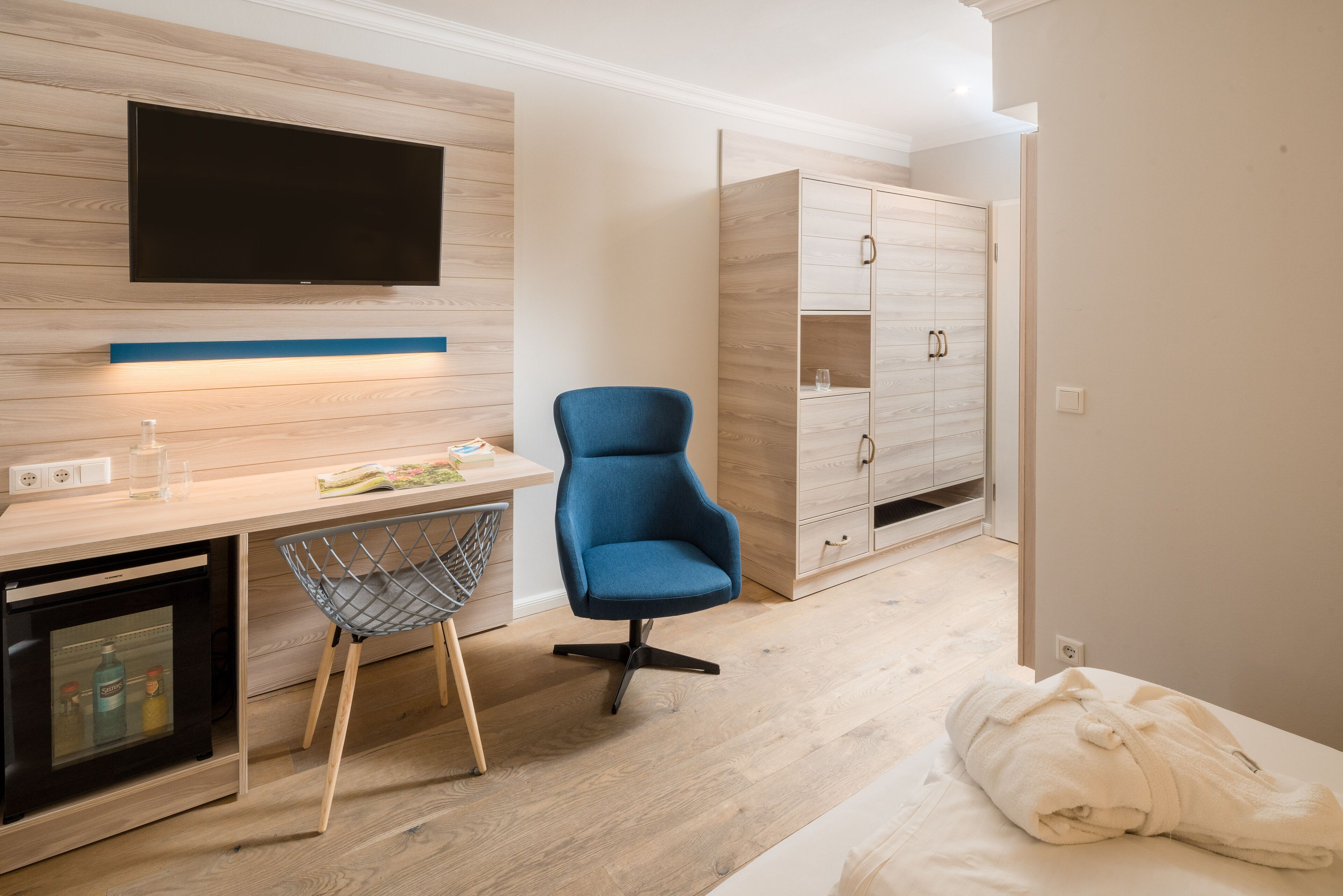 Een moderne kamer met een houten vloer, een bureau met een flatscreen-televisie, een blauwe stoel, een kledingkast en een bed met een witte deken.