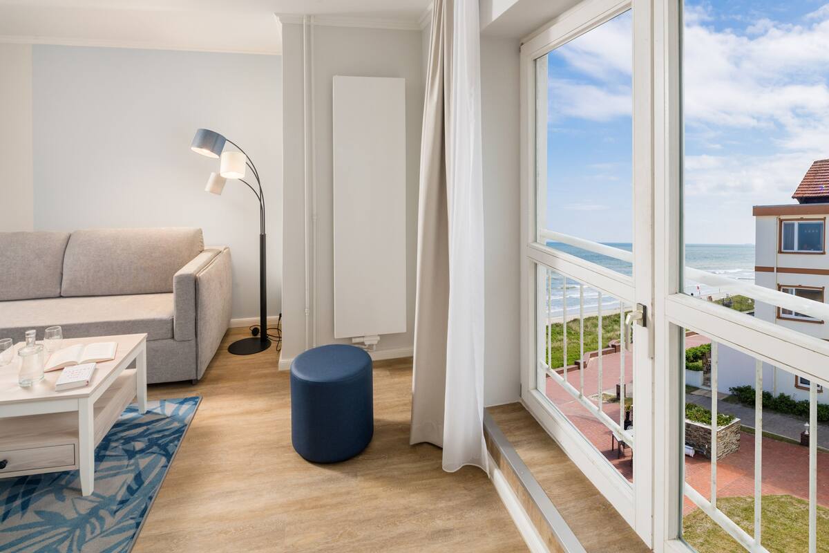 A modern living room with a sofa, coffee table, and floor lamp. A large window offers a view of the ocean and a building across the street.