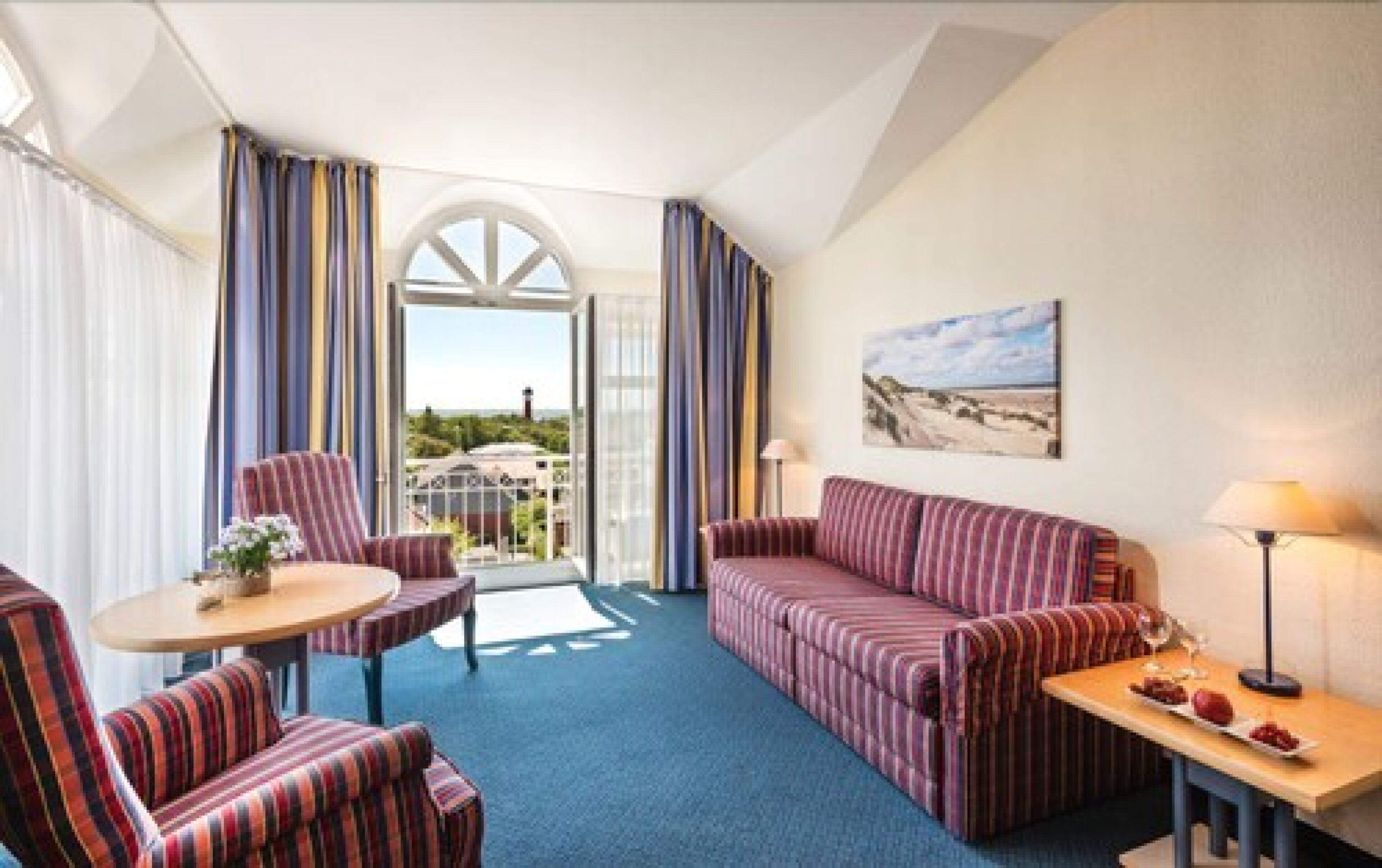 A hotel room with a sofa, armchair, and a small table with a lamp. There is a large window with curtains, and a painting on the wall.
