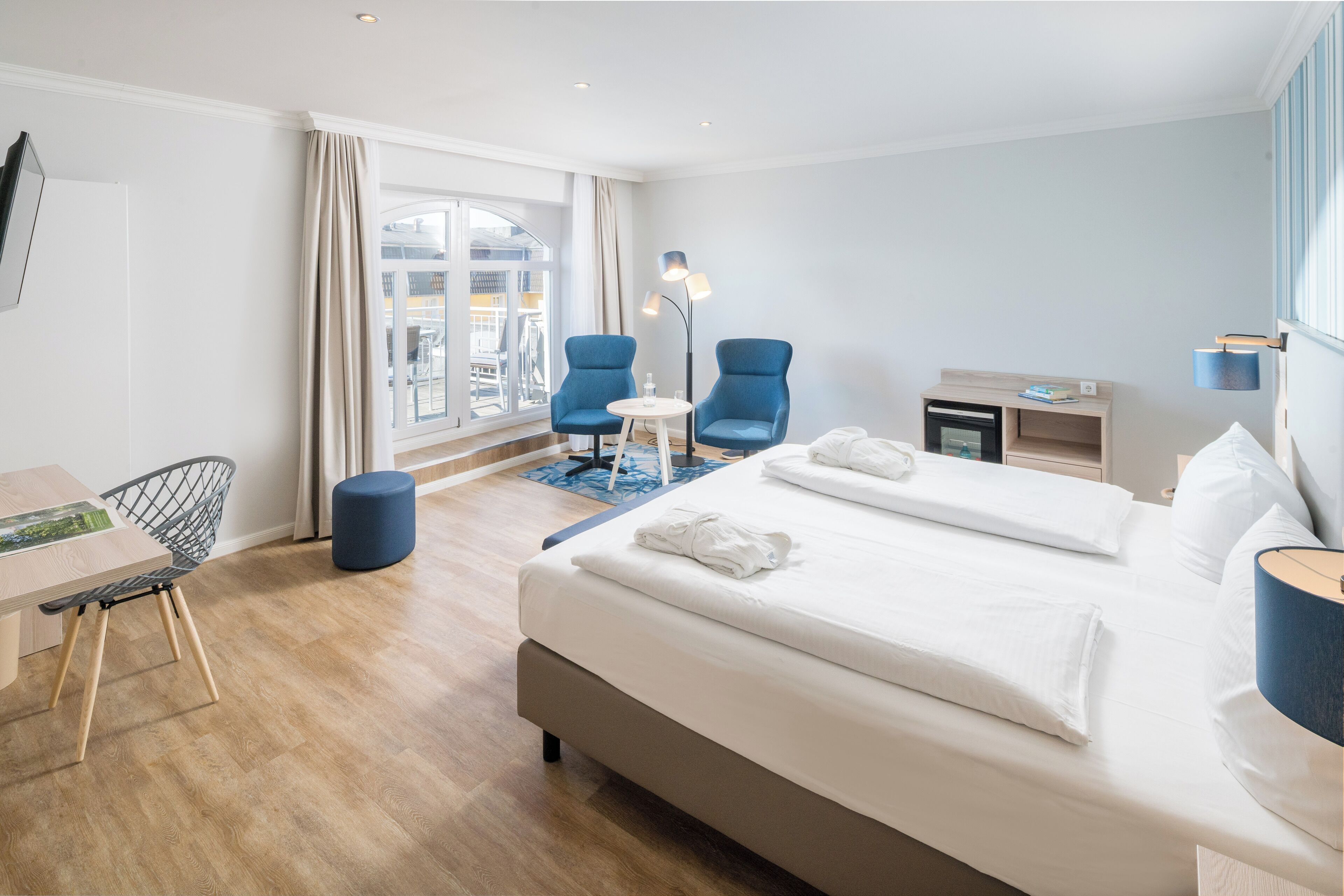 A modern hotel room with a large bed, two blue armchairs, a small table, a desk, and a view of the city through large windows.