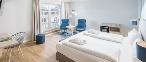 A modern hotel room with a large bed, two blue armchairs, a small table, a desk, and a view of the city through large windows.