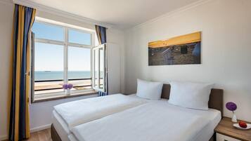 A hotel room with a large bed, a window with curtains, a painting of a beach scene, and a small vase with flowers.