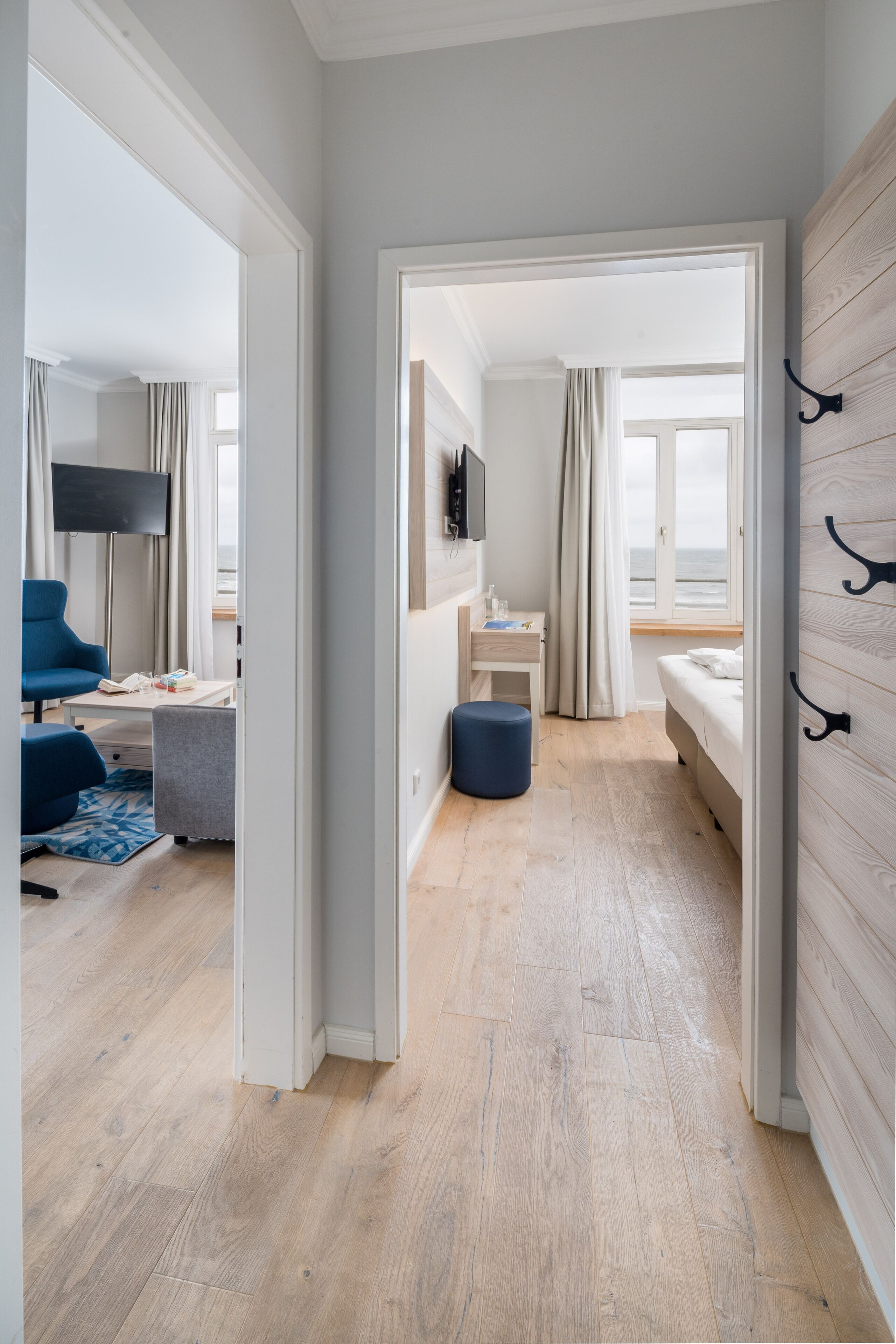 A modern hotel room with a wooden floor, a bed, a desk with a chair, a TV mounted on the wall, and a view of the sea through the window.