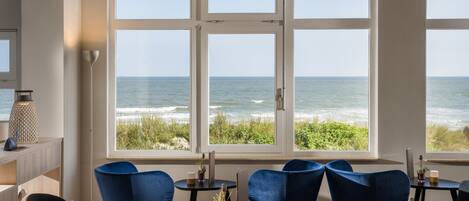 A room with a large window overlooking the ocean, featuring a round table and blue chairs.