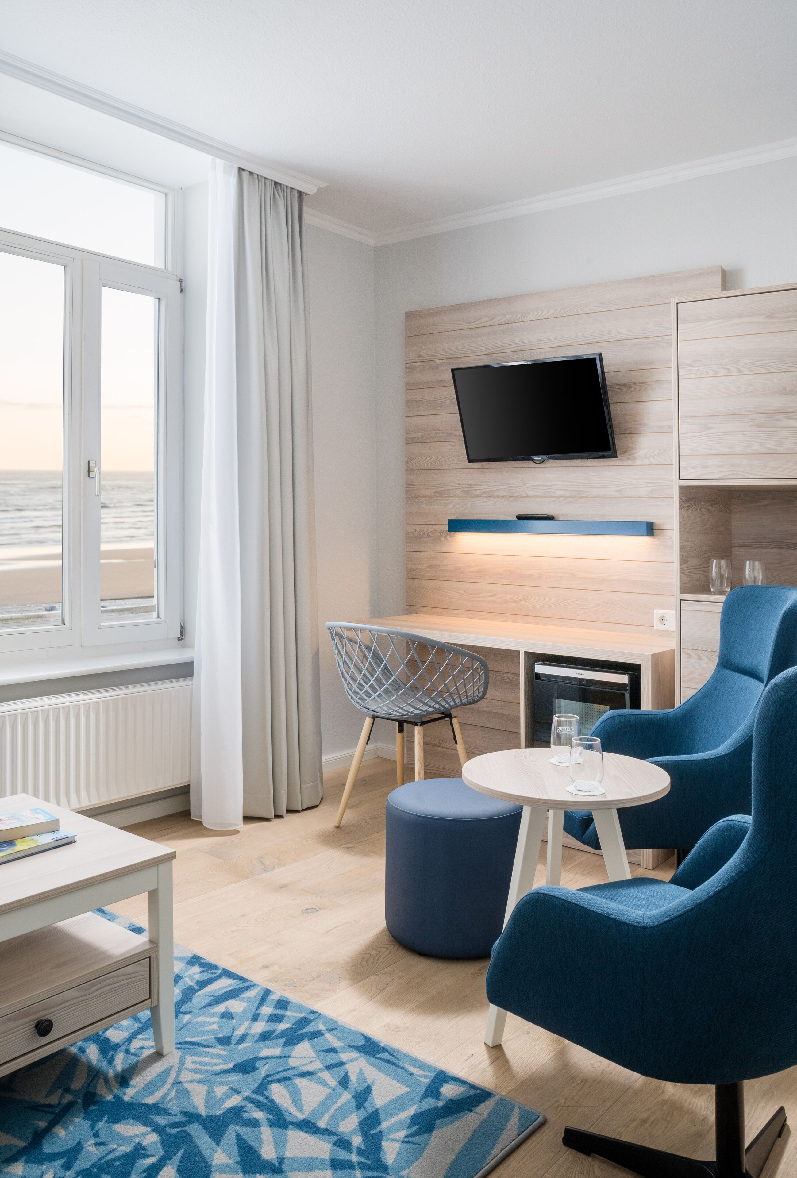 A modern hotel room with a large window, a flat-screen TV, a wooden desk, two blue armchairs, and a small round table.