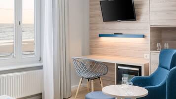 A modern hotel room with a large window, a flat-screen TV, a wooden desk, two blue armchairs, and a small round table.