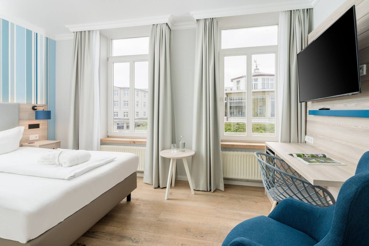 A hotel room with a bed, a desk, a chair, a TV, and large windows with curtains.