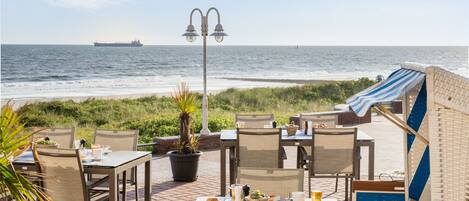 An outdoor dining area with tables and chairs, a potted plant, and a view of the ocean with a ship in the distance.