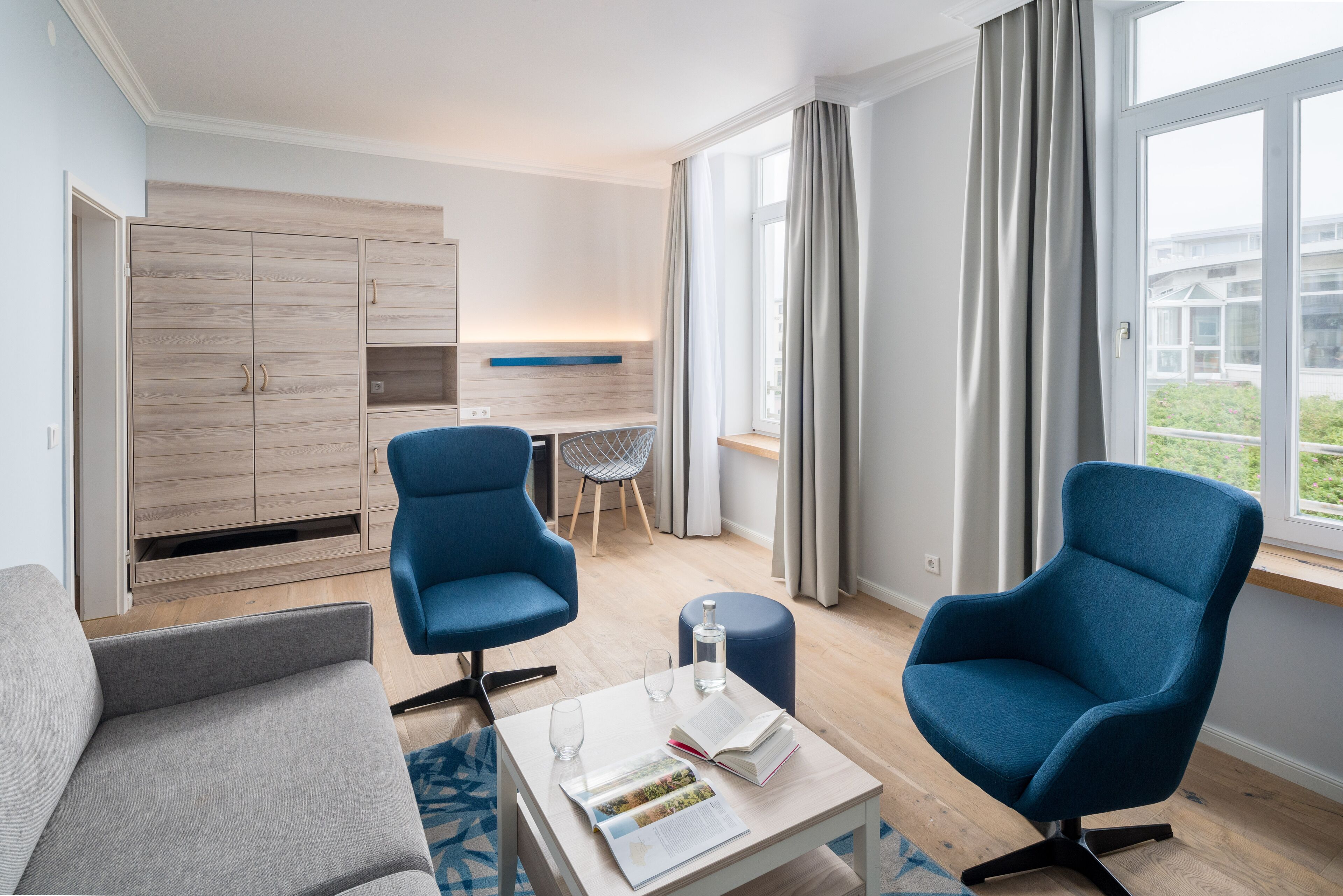 Ein modernes Hotelzimmer mit einer Couch, zwei Sesseln, einem kleinen Tisch mit Buch und Glas, einer Minibar und Blick ins Freie.