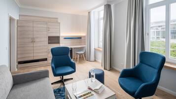 Ein modernes Hotelzimmer mit einer Couch, zwei Sesseln, einem kleinen Tisch mit Buch und Glas, einer Minibar und Blick ins Freie.