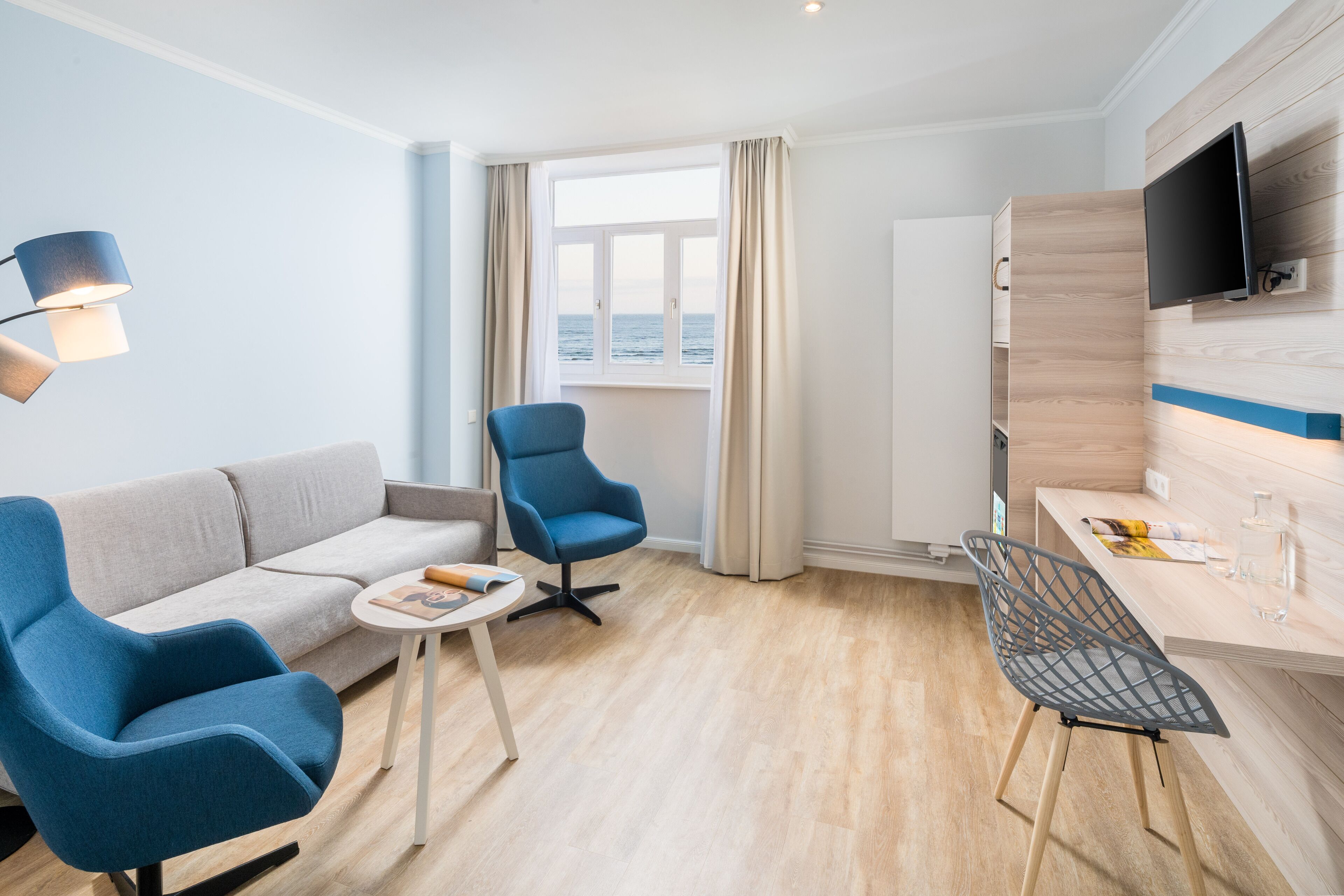 A modern hotel room with a sofa, two chairs, a small table, a TV, and a window with a view of the sea.