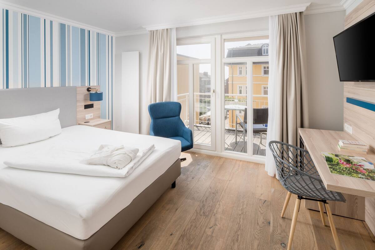 A hotel room with a bed, a desk, a chair, a TV, and a balcony with a view of buildings.
