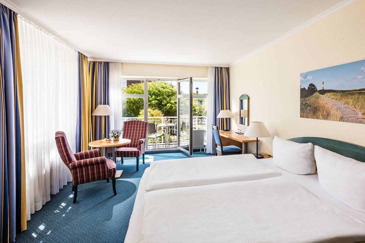 A hotel room with a large bed, a desk with a chair, a small table with a chair, and a balcony with a view of greenery.