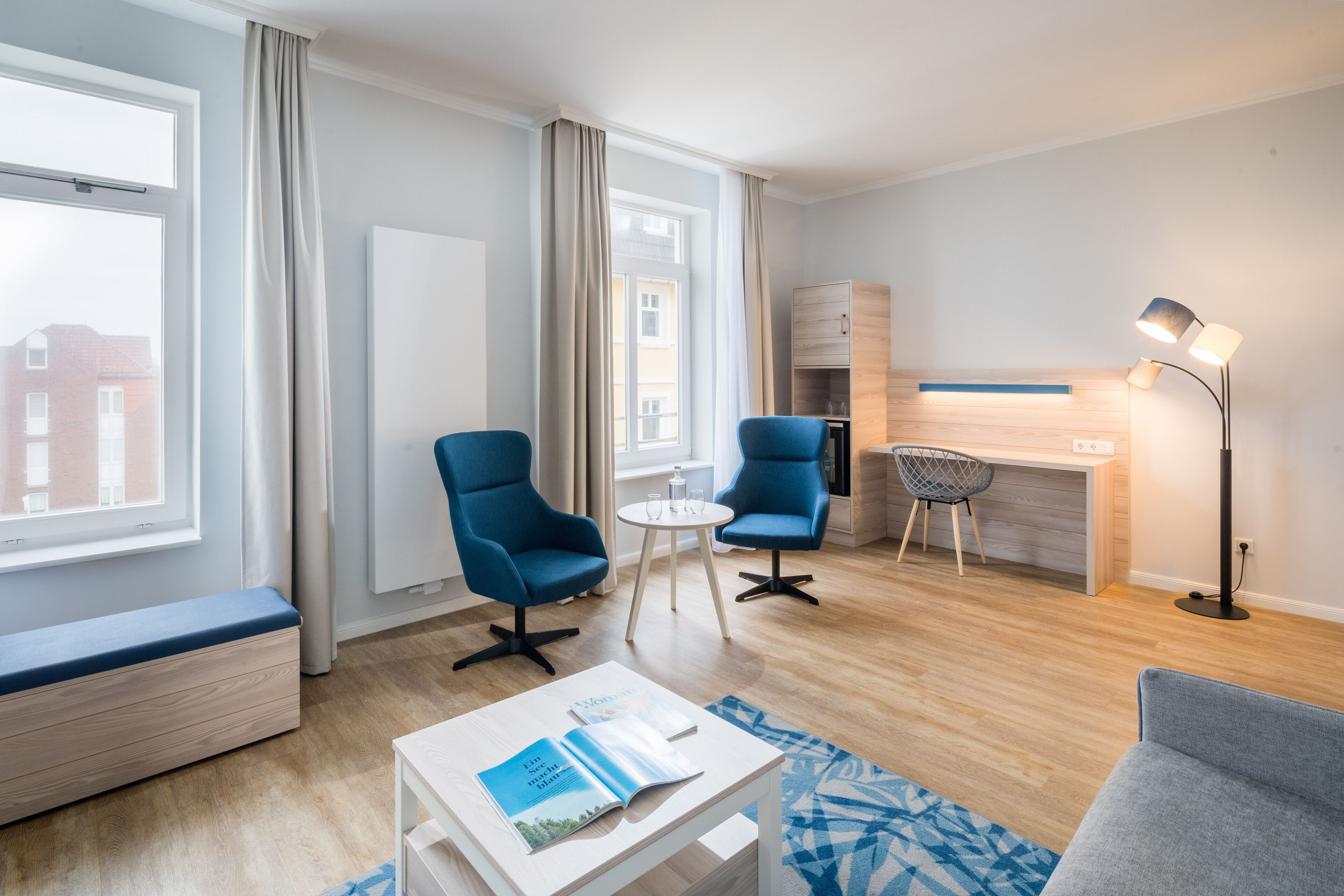 A modern hotel room with a wooden floor, a desk, two blue armchairs, a small round table, a white coffee table with magazines, a blue patterned rug, and a floor lamp.