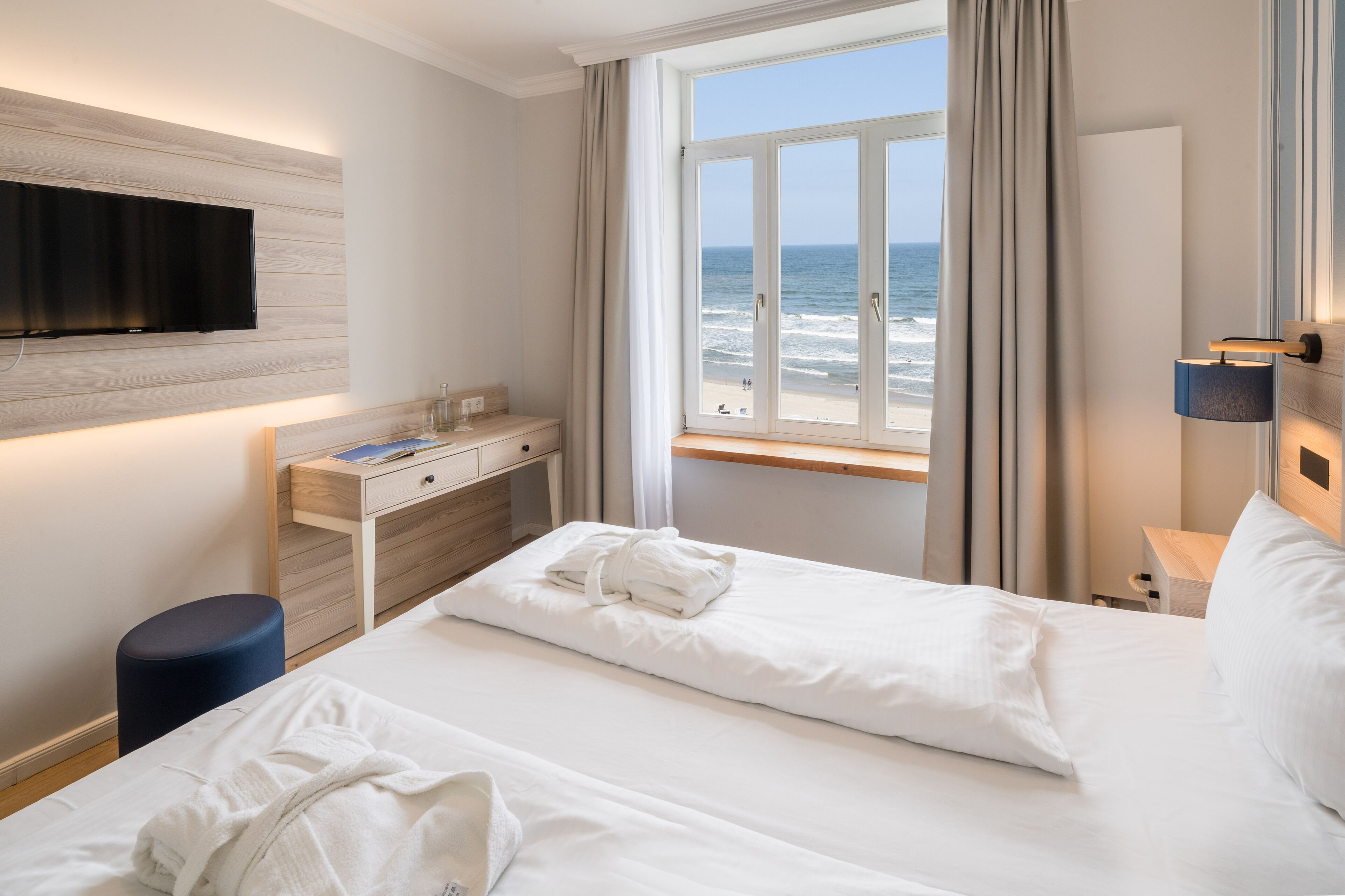 A hotel room with a bed, a desk, a TV mounted on the wall, and a window with a view of the ocean.