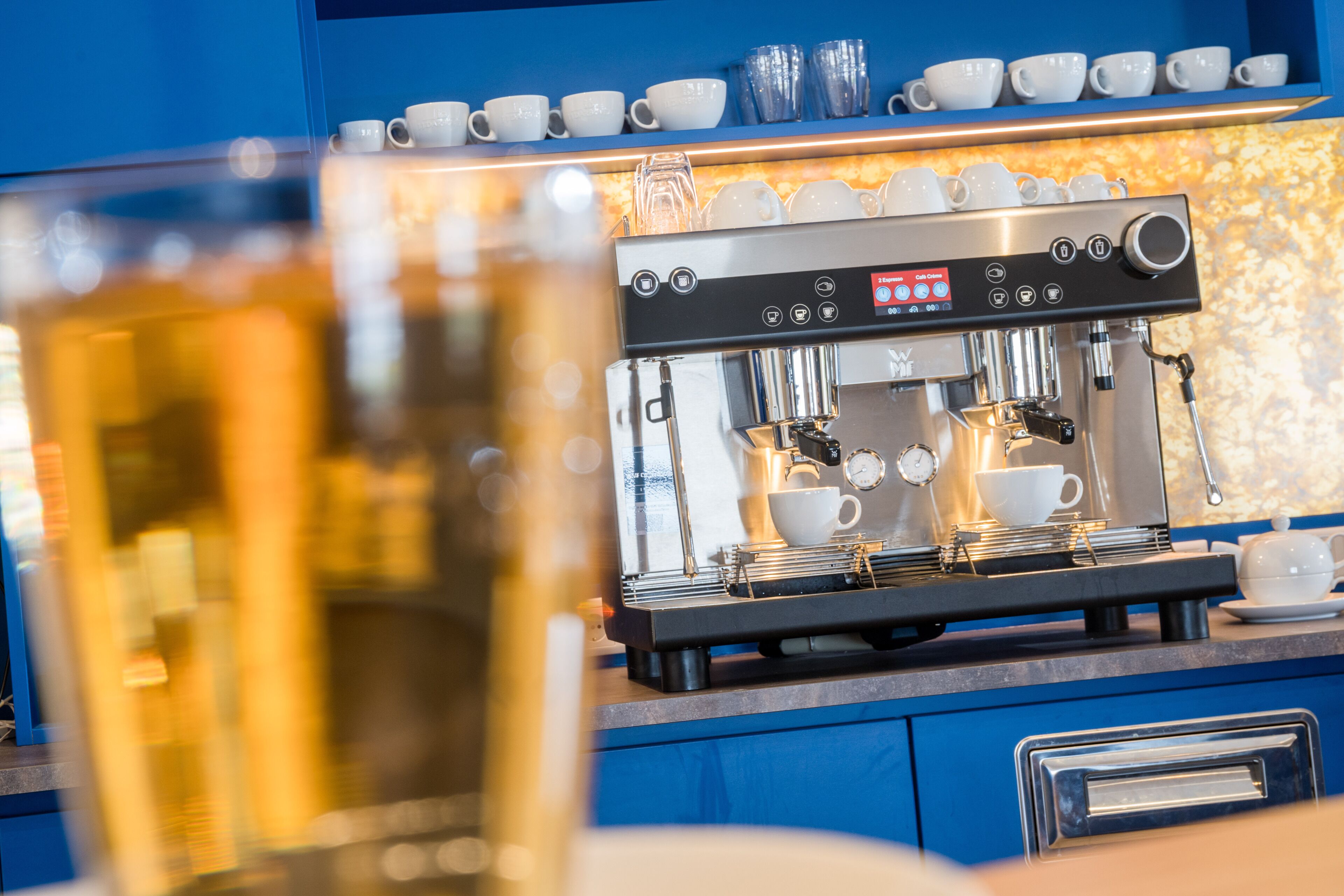 A coffee machine with multiple cups and a clock, set against a blue wall with shelves of cups.