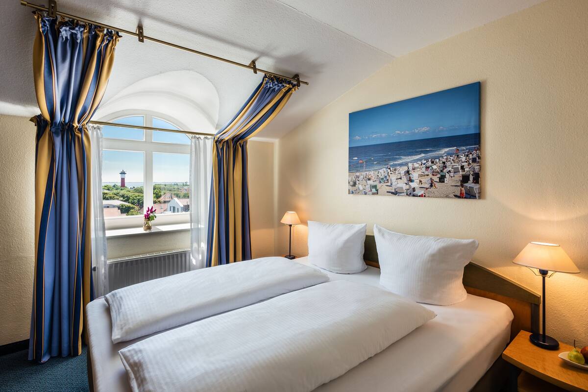 A hotel room with a large bed, two bedside lamps, a painting of a beach scene, and a view of a lighthouse through the window.