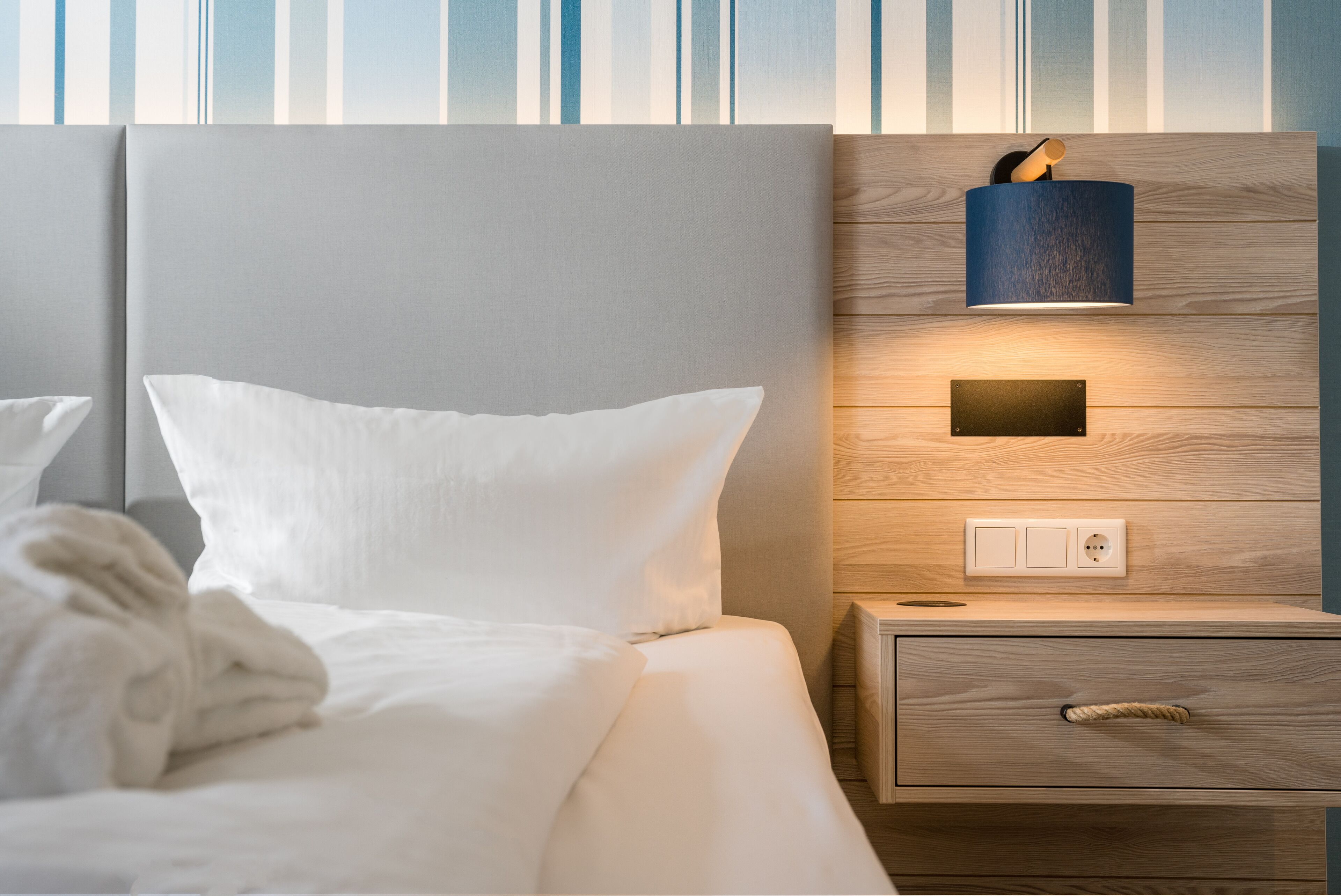Ein ordentlich bezogenes Bett mit weißen Leinen und Kissen, ein Nachttisch aus Holz mit einer Lampe und ein an der Wand befestigter Lichtschalter.