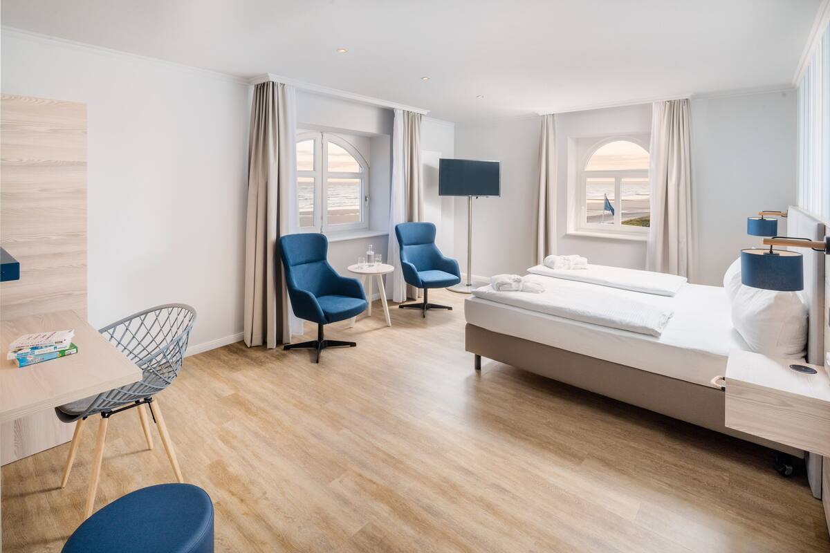 A hotel room with a bed, two blue armchairs, a small table, a TV, and a window with a view of the beach.