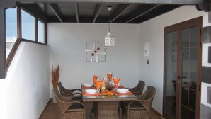 Un coin repas avec une table dressée pour quatre personnes, entourée de chaises en osier, sous un plafond avec des poutres apparentes.