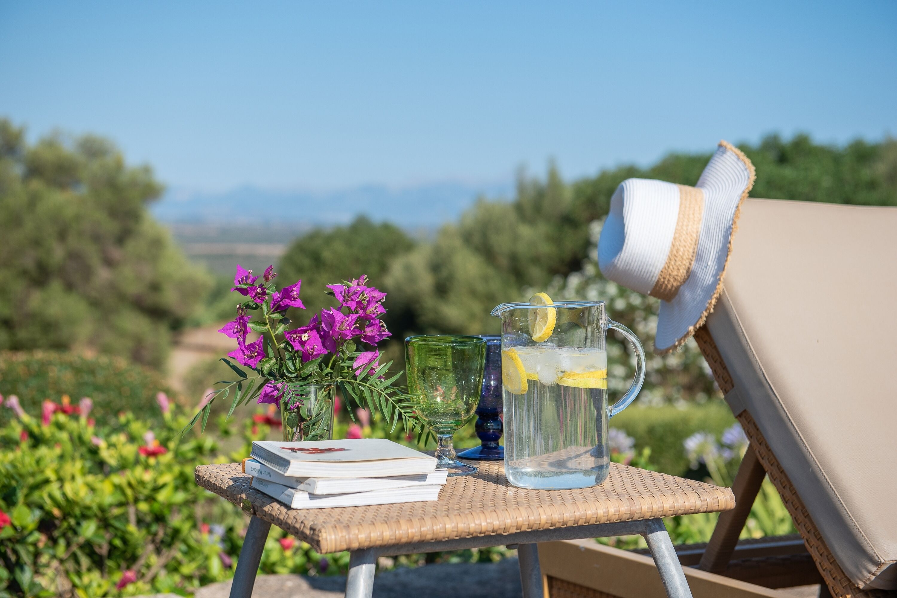 A wicker table with a pitcher of water, a glass, and books, set outdoors with a view of greenery and mountains.